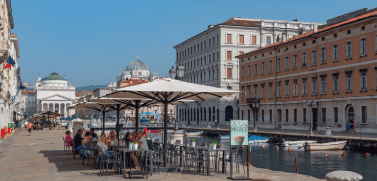 Tavoli di un locale, affacciato sul Canal Grande di Trieste, in cui si mangia all'aperto. Si vede la chiesa di Sant'Antonio Nuovo. La foto è stata scattata di giorno.