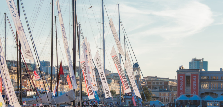 Imbarcazioni a vela ormeggiate nel golfo di Trieste, pronte a partecipare alla regata Barcolana, con la folla sulle banchine, i palazzi storici della città e gli stand sullo sfondo.